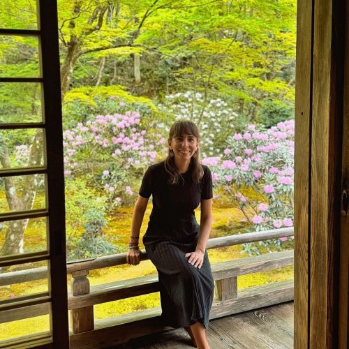 Diana Chiu Baptista, fundadora da Macro Viagens. de vestido preto, sentada de costas para um jardim japonês muito verde e com algumas flores