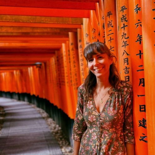 Diana, fundadora da Macro Viagens, sorri para a fotografia, vestida com um vestido verde com flores, num cenário com tori vermelhos, no Japão.