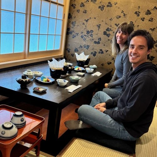 Diana e Igor fundadors da Macor Viagens, sentados num restaurante típico japonês com chão em tatami e mesas baixinhas sorriem para a fotografia antes de começarem a comer (a comida esta servida em pequenas taças)