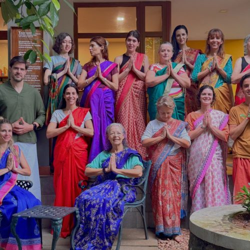 Grupo de ocidentais vestidos com os coloridos trajes típicos de kerala a fazer pose para uma fotografia, com as mãos em "namasté", num centro ayurveda na índia onde fizeram um retiro em grupo