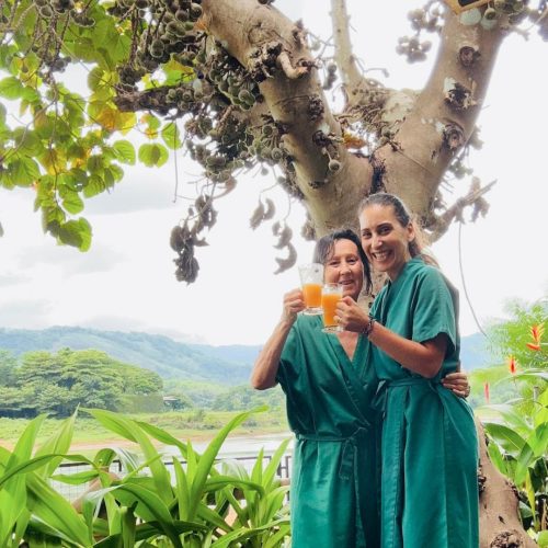 Duas mulheres vestidas com robes para tratamentos ayuvreda fazem um brinde sorridentes com dois sumos ayurvédicos, estão num cenário natural, em kerala