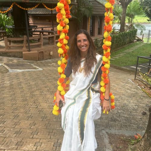Mulher sorridente vestida de saree tradicional de Kerala está sentada num baloiço decorado com flores, ao ar livre, num cenário natural do sul da Índia