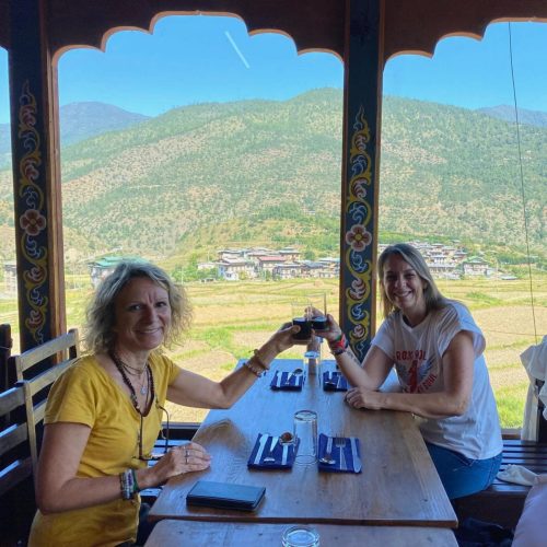 Duas mulheres viajantes brindam num restaurante no Butão, junto a uma janela com uma vista para um vale verdejante