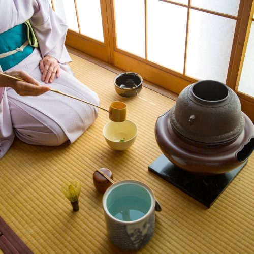 Senhora japonesa ajoelhada, com kimono rosa claro, a servir cá num chão em tatami