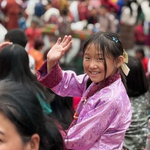 Uma menina butanesa acena sorridente, no meio da população. Está vestida com o traje tradicional, de blusa cor-de-rosa, no meio da população local.