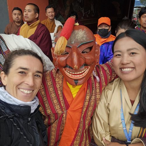 Uma viajante da macro Viagens, um participante de uma dança Chamm - mascarado - e uma guia butanesa tiram uma selfie.