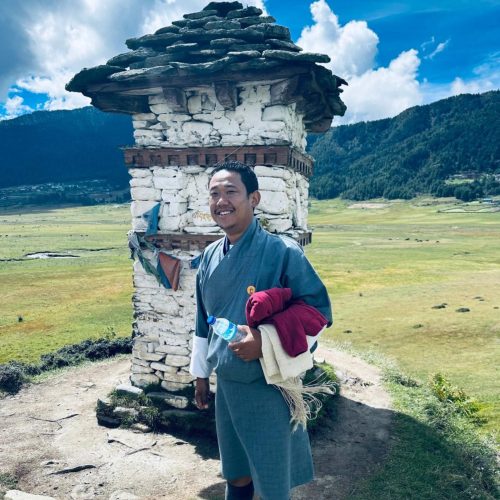 Um homem butanês sorridente, vestido com a roupa tradicional de costas para um pequeno monumento no Butão