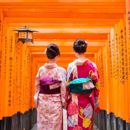 Duas mulheres japonesas, com os trajes tradicionais, caminham de costas para a fotografia, por vários tori vermelhos