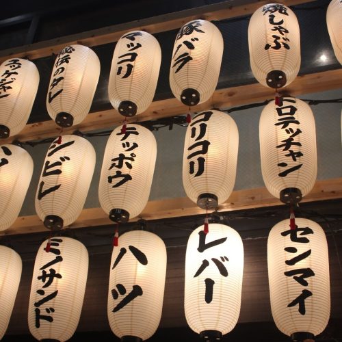 Várias lapadas de papel, brancas com caracteres japoneses em preto. Estão iluminadas e é de noite.