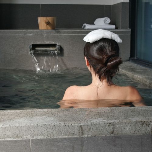 Mulher de costas sentada num Onsen, o banho termal japonês. Tem uma pequena toalha branca pousada sobre a cabeça.