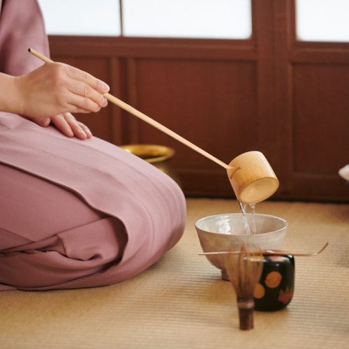 Mulher japonesa ajoelhada, com kimono rosa claro, só se vê as pernas e a mão, está a servir um chá